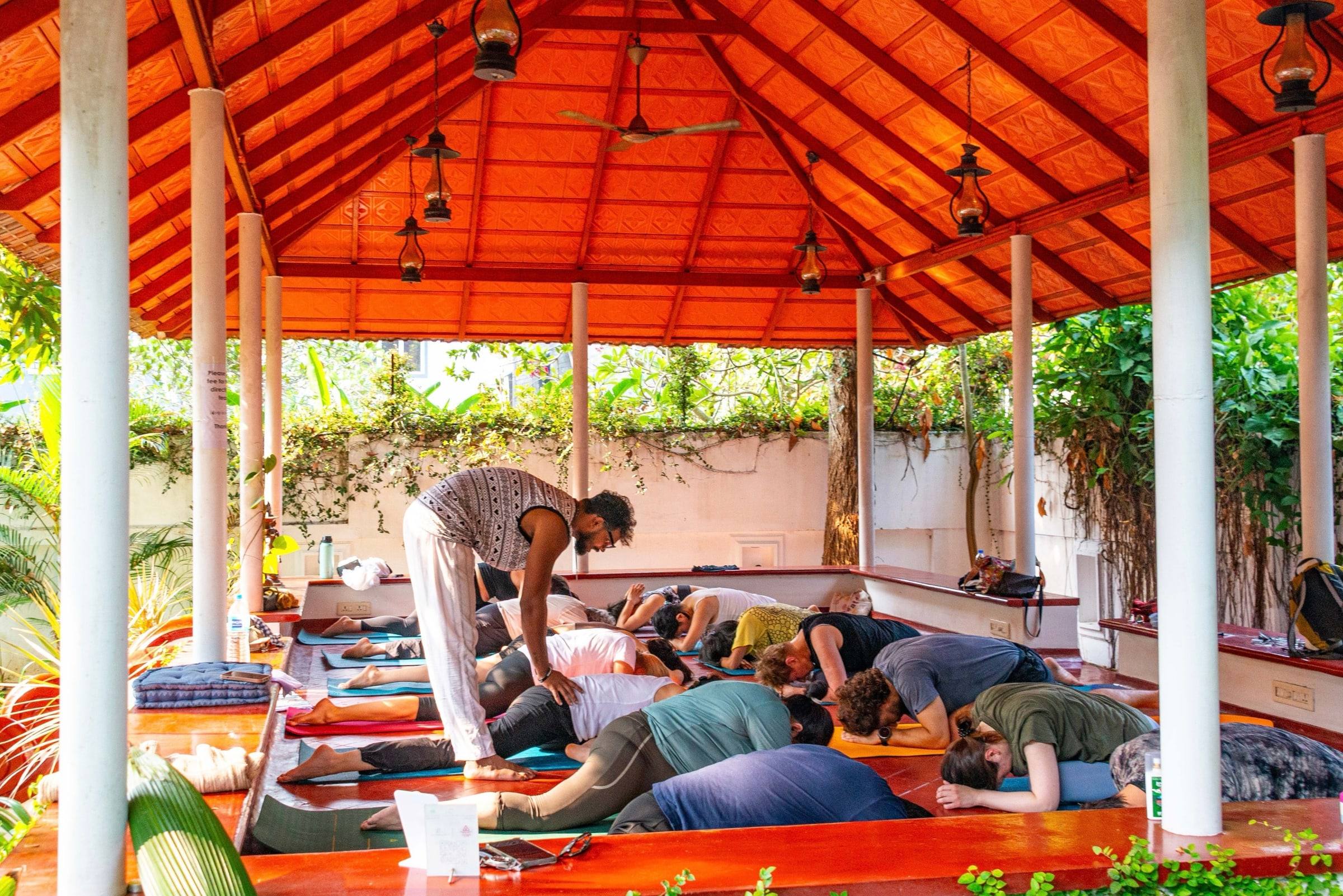 The teacher moving between students resting in child’s pose under the orange pavilion roof