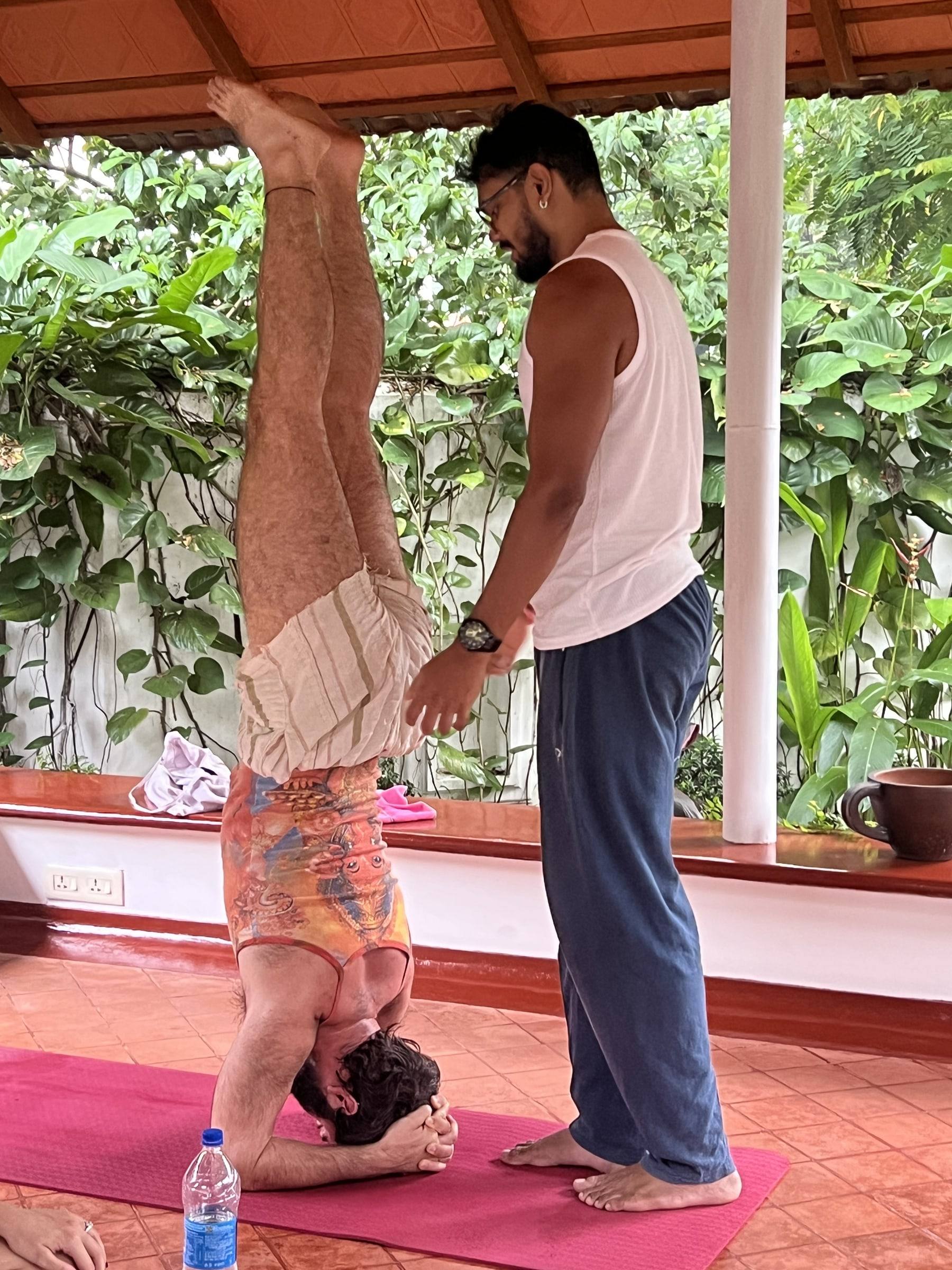 The teacher spotting a student’s headstand in the pavilion, garden greenery behind