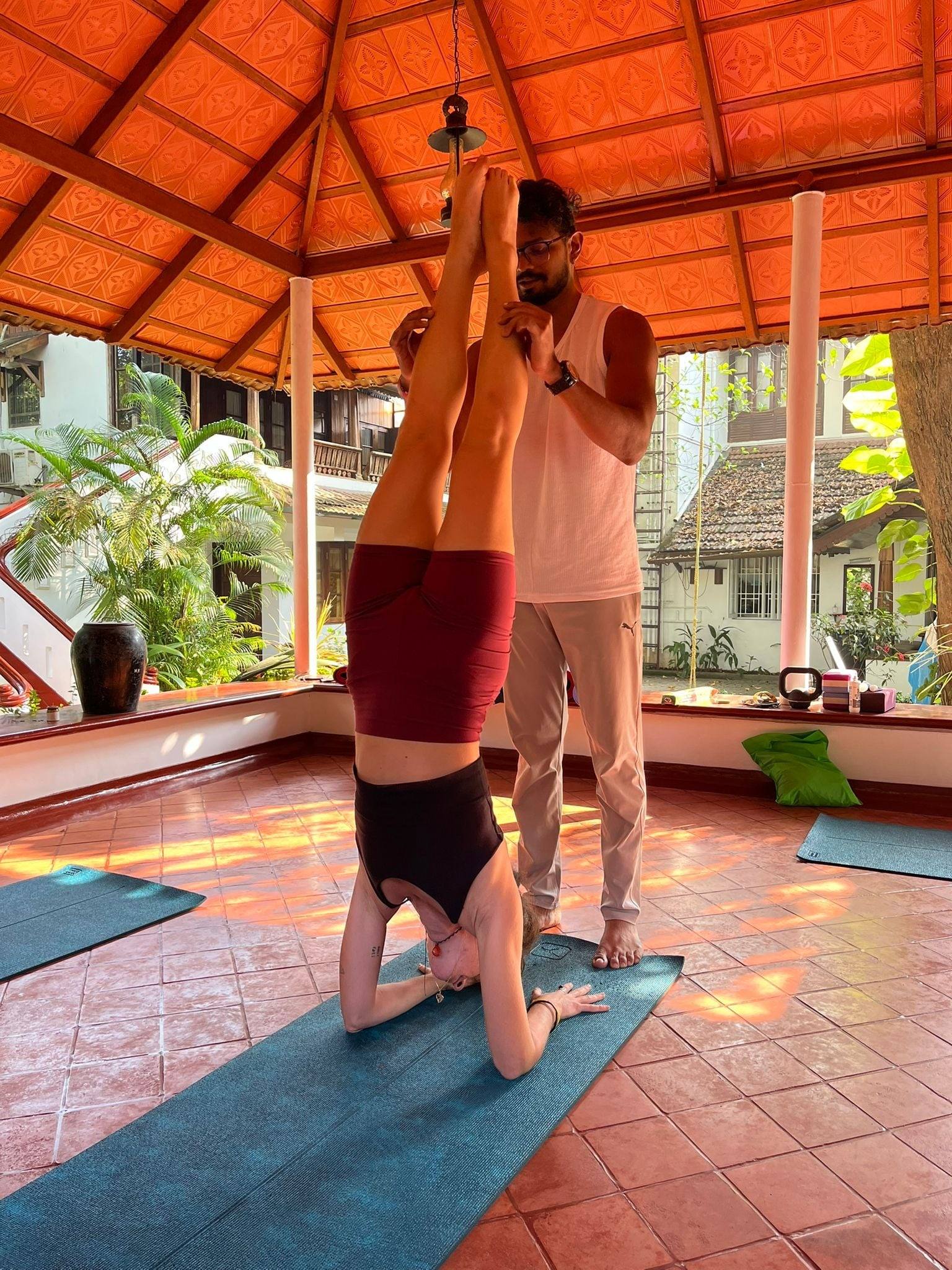A student in headstand with the teacher steadying her legs under the tiled pavilion roof
