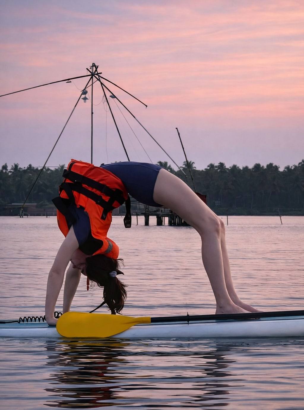 A wheel pose on a paddleboard at dusk, with a Chinese fishing net rising behind on the water